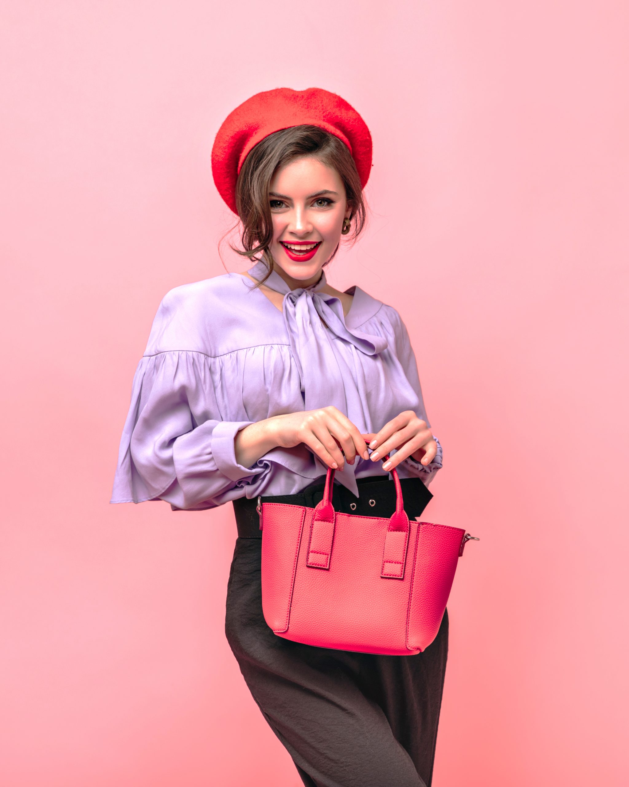 Flirtatious woman in beret, blouse and trousers holding red bag on pink background.